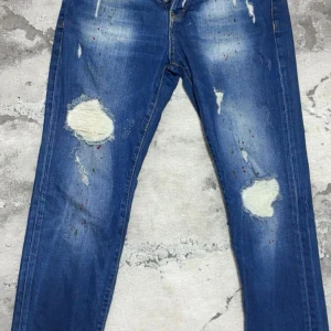 Blå Replay jeans med slitningar - Säljer ett par blå Replay jeans med coola slitningar och färgstänk på framsidan. Jeansen har klassisk femficksdesign, grönt Replay-märke bak och raka ben. Materialet är jeans med en skön passform som funkar till streetwear. Perfekt för dig som gillar en edgy stil.