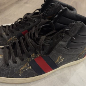 Gucci sneakers med tigerprint - Snygga high-top sneakers från Gucci med mörkgrå tyg och klassiska röda och marinblå ränder på sidan. Skorna har coolt tigerprint och GG-mönster över hela ytan. Baksidan har en röd och en blå häl med Gucci-logga. Svarta snören och vadderad krage för extra komfort. Ni välkomna och lägga era bud (OBS: skor är äkta)