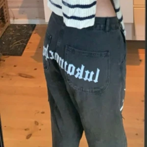 Svarta jeans med tryck från lukamachain - Unika svarta jeans från lukamachain med stort vitt tryck över bakfickorna och ett silvrigt tribal-motiv på ena benet. Modellen är lågmidjad med raka ben ch riktigt baggy! Jeansen är tillverkade i denim och har knapp och dragkedja i midjan. Dessa jeans säljs inte längre och är ett riktigt kap!