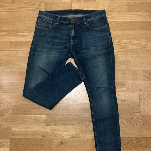 Blå jeans från Nudie Jeans Co - Snygga blå jeans från Nudie Jeans Co med klassisk femficksdesign och orangea sömmar. Jeansen har en rak passform och är tillverkade i slitstarkt denim med coola tvättade detaljer. Perfekt för dig som gillar stilrena och tidlösa jeans.