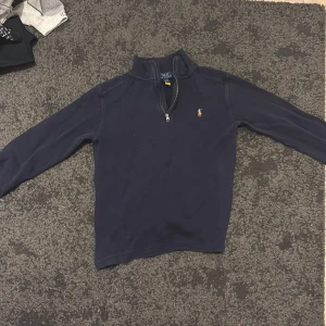 Marinblå half zip tröja Polo Ralph Lauren - Marinblå långärmad tröja från Polo Ralph Lauren med half zip och klassisk ståkrage. Tröjan har det ikoniska broderade Polo-logot på bröstet och är gjord i mjuk bomull. Perfekt för dig som gillar stilrena och tidlösa plagg.