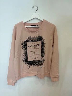 Beige sweatshirt med tryck från KappAhl - Snygg beige sweatshirt från KappAhl med svart tryck framtill där det står 'Magnifique' och franska texter. Tröjan har rund hals, långa ärmar och ribbade muddar vid ärmslut och nederkant. Perfekt för dig som gillar coola prints och fransk vibe.