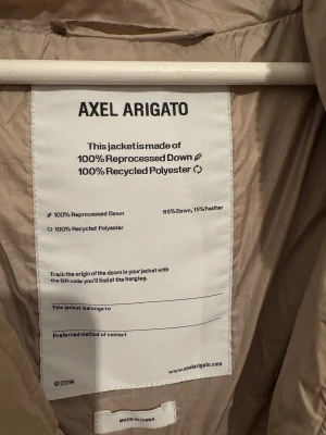 Beige pufferjacka från Axel Arigato - Säljer en beige pufferjacka från Axel Arigato med huva och snörning i midjan. Jackan har dragkedja framtill och tryckt logga på bröstet. Tillverkad av 100% återvunnen polyester och reprocessad dun. Perfekt för kalla dagar och har en clean, modern look. Pris kan diskuteras.