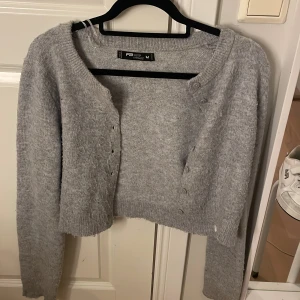 Grå croppad kofta från FB Sister - Grå stickad kofta från FB Sister i croppad modell med långa ärmar och rund hals. Koftan har knappar framtill och ribbade muddar vid ärmslut och nederkant. Perfekt för dig som gillar en chill och enkel stil.