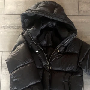 Svart pufferjacka med huva - Säljer en svart pufferjacka med stor, mysig huva och dragkedja framtill. Jackan är i bra skick och använd få gånger från Dressy. Pris kan diskuteras!