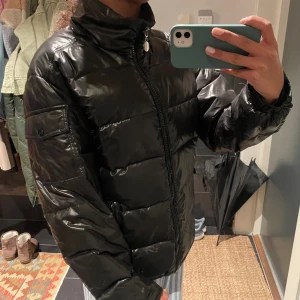 Svart pufferjacka med glansig yta - Svart pufferjacka med glansig finish och hög krage. Jackan har långa ärmar, två fickor med knappstängning och dragkedja framtill. Perfekt för kalla dagar och har en modern, clean look som funkar till streetwear.