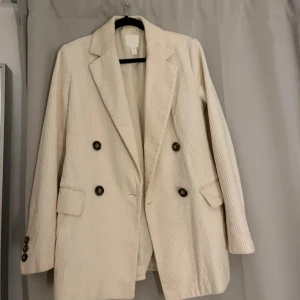 Vit manchesterkavaj med knappar - Snygg vit kavaj i manchester från H&M med dubbelknäppning, Kavajen har fyra mörka knappar framtill, fickor med lock och knappar vid ärmslut. Passar dig som gillar stilrena och trendiga plagg med lite retrovibe.