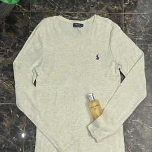 Ljusgrå långärmad tröja Polo Ralph Lauren - Snygg ljusgrå långärmad tröja från Polo Ralph Lauren med klassisk rund hals och lila broderad logga på bröstet. Tröjan är i mjuk bomull och har en clean, enkel look som funkar till jeans eller shorts. Perfekt för dig som gillar stilrena basics.
