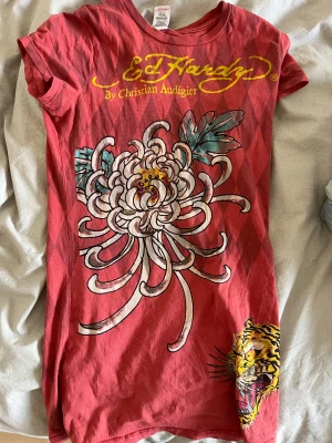 Röd Ed Hardy t-shirt med tryck - Säljer en röd t-shirt från Ed Hardy med coolt blommönster och tigertryck framtill samt en stor cowboy-figur på ryggen. T-shirten har korta ärmar, diamantmönster i bakgrunden och gula logotyper. Materialet är mjuk bomull och passformen är normal.