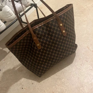 Brun shoppingväska  - Snygg shoppingväska  i brunt med klassiskt monogrammönster och långa handtag i skinn. Väskan är rymlig och perfekt för dig som vill ha plats för allt. Guldiga detaljer vid handtagen ger en lyxig känsla.