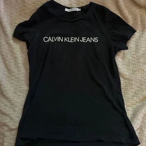 Svart t-shirt från Calvin Klein Jeans - Svart t-shirt från Calvin Klein Jeans med vit logotyp tryckt över bröstet. Klassisk rund hals och korta ärmar. Tillverkad i mjuk bomull som känns skön mot huden. Perfekt för en enkel och stilren look.