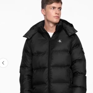Svart pufferjacka från Calvin Klein - Svart pufferjacka från Calvin Klein Jeans med ikoniska CK-loggan på bröstet och ärmen. Jackan har huva, dragkedja framtill och två sidofickor med dragkedja. Perfekt för kalla dagar och har en clean, modern look som funkar till jeans och sneakers.! INTE FAST PRIS SÅ GE ERBJUDANDE!