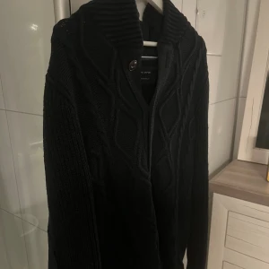 Svart stickad kofta från Marc O'Polo - En svart, grovstickad kofta från Marc O'Polo med snyggt flätmönster och ribbad krage. Koftan har knappar framtill och långa ärmar med förstärkta armbågslappar. Perfekt för dig som gillar klassisk stil med en modern twist. Prisen kan förhandlas! Ny pris: 2199 kr autentisk modell.