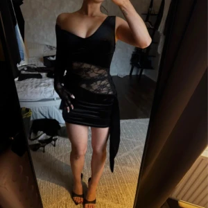 Svart SNYGG spetsklänning - Säljer en svart kortklänning med asymmetrisk design och v-ringad hals. Ena sidan har lång spetsärm och transparent spets över midjan, medan andra sidan är ärmlös. Klänningen har en snygg knyt-detalj i midjan och sitter tight. Perfekt för dig som vill sticka ut med en unik look.