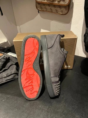 Christian Louboutin grå sneakers med nitar - Säljer ett par grå sneakers från Christian Louboutin med coola nitar på tån och klassisk röd sula. Skorna har snörning och är tillverkade i mocka med rund tå och platt sula. Perfekt för dig som vill sticka ut med en edgy vibe. Använda 1 gång 2 spikar borta längst fram på skon men kommer sätta på 2 nya imorgon så det kommer sitta på när du får skorna.