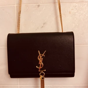 Svart YSL axelväska med guldkedja - Snygg svart axelväska från YSL med guldfärgad kedja och ikonisk logga framtill. Väskan har en stilren, rektangulär form och är tillverkad i slätt skinn. Framsidan pryds av en guldfärgad tofsdetalj som ger extra lyxig vibe. Perfekt för dig som gillar exklusiva accessoarer.