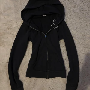 Svart hoodie från Hollister med dragkedja - Svart hoodie från Hollister med vit broderad logga på bröstet och texten 'California 1922'. Tröjan har huva, långa ärmar, två fickor framtill och hel dragkedja. Mjuk och skön i bomullsmaterial, perfekt för chill dagar.