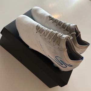 Vita fotbollsskor Skechers Razor - Säljer ett par vita Skechers Razor fotbollsskor med blå detaljer och snörning. Skorna har en smal och sportig siluett med rund tå och platt sula med blå dobbar för bra grepp på planen. Dem är knappt andvända så är i bra skick. 