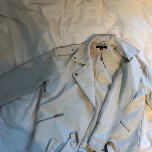 Vit / begei skinnjacka från Missguided - Säljer en snygg vit - begie skinnjacka från Missguided med klassisk bikerstil. Jackan har silvriga dragkedjor, knappar och ett bälte i midjan. Modellen är kortare och har långa ärmar!