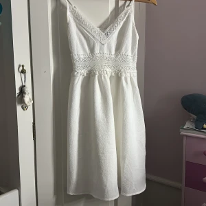Vit spetsklänning med smala axelband - Superfin vit klänning med smala axelband och v-ringning. Klänningen har spetsdetaljer längs urringningen och midjan, samt en lätt rynkad kjol. Materialet är strukturerat och känns luftigt, perfekt för varma dagar. Söt och romantisk stil med broderade blommor i spetsen. Använt 2 gånger, skit gullig och fin ☺️ säljer för den inte kommer till användning ☺️❤️