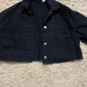 Svart croppad overshirt XS från H&M - Svart croppad overshirt från H&M i storlek XS. Jackan har två stora fickor framtill, silverfärgade knappar och rå kant nedtill för en chill vibe. Perfekt att slänga över en t-shirt för en avslappnad look.