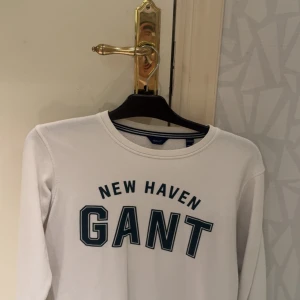 Vit långärmad sweatshirt från GANT - Vit sweatshirt från GANT med trycket 'NEW HAVEN GANT' i mörkblått på bröstet. Tröjan har rund hals, långa ärmar och är tillverkad i mjuk bomull med fleece på insidan. Perfekt för dig som gillar klassisk och sportig stil.