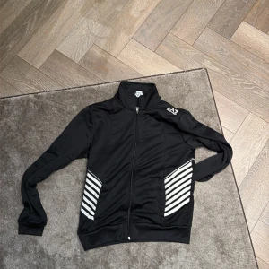 Svart EAZ vindjacka med vita ränder - Svart hoodie från EA7 med vita diagonala ränder på sidorna och en vit logga på axeln. Tröjan har hel dragkedja framtill, ståkrage och långärmad design. Materialet är mjukt och syntetiskt, perfekt för en sportig look.