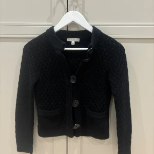 Svart stickad kofta från Dagmar - Svart stickad kofta från Dagmar med snyggt strukturmönster och stora knappar framtill. Koftan har två fickor och långärmade ärmar. Perfekt för dig som gillar klassisk stil med en twist. Koftan är använd fåtals gånger och är i ett mycket bra skick💕
