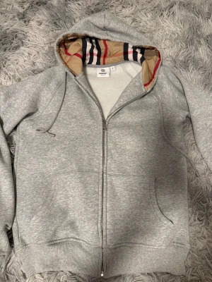 Grå hoodie från Burberry med dragkedja - Säljer en grå hoodie från Burberry, det står storlek M men sitter exakt som XS. Tröjan har klassisk Burberry-rutig huva, snörning med metalländar och en snygg dragkedja framtill. Mjuk bomullskänsla och två fickor fram. Perfekt för dig som gillar stilrena och lyxiga detaljer.