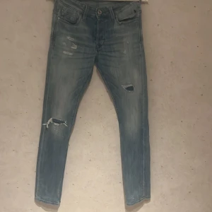 Slim Glenn jeans från Jack & Jones - Snygga ljusblå jeans från Jack & Jones, modell Slim Glenn. De har slitna detaljer och hål vid knäna för en cool, avslappnad vibe. Klassisk femficksdesign och smal passform som sitter tajt längs benen. Perfekta för dig som gillar streetstyle.