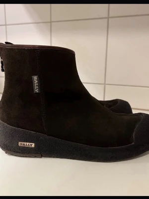Bruna mockaboots från Bally - Säljer ett par bruna boots från Bally i mjuk mocka med grov sula och dragkedja bak. Skorna har en stilren och minimalistisk design med rund tå och diskret Bally-logga på sidan och sulan. Perfekta för dig som gillar klassiska men coola skor. Det har släppt lite på ena skon och jag kan sända bild vid begäran  dem är fint använt Swish är det som gäller 🌺 väl mött