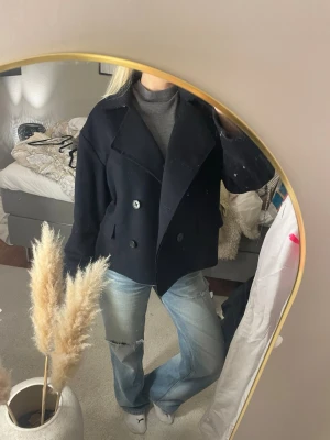 Svart/marinblå kort kappa i ull - Säljer en kort kappa med dubbelknäppning och bred krage. Jackan är i ullmaterial och har en loose passform som ger en chill vibe. Perfekt att slänga på över en hoodie eller stickad tröja. Klassisk look med stora knappar och fickor framtill. Nypris 2700 kr 
