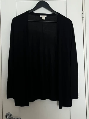 Svart kofta från H&M Basic, stl M - En enkel svart kofta från H&M Basic i storlek M. Koftan har lång ärm, öppen framtill och är gjord i ett tunt och mjukt material som känns skönt mot huden. Perfekt att slänga över en t-shirt eller linne när du vill ha en chill och stilren look.
