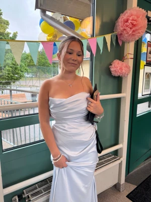 Ljusblå axelbandslös långklänning - Säljer en elegant ljusblå långklänning med axelbandslös design och draperad överdel. Klänningen har en snygg omlottkänsla och är tillverkad i ett glansigt satinliknande material som ger en lyxig känsla. Perfekt för bal eller student!