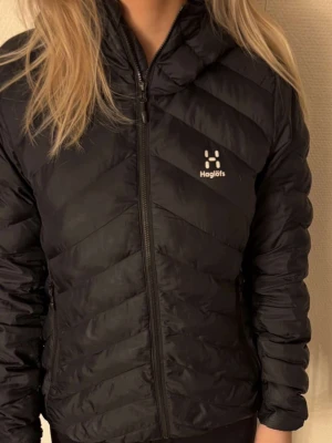 Svart pufferjacka från Haglöfs dam S - Svart pufferjacka från Haglöfs i dammodell, storlek S. Jackan har huva, dragkedja framtill och två sidofickor med dragkedja. Snyggt quiltad design med diskret logga på bröstet. Perfekt för kalla dagar och har en modern, sportig look.