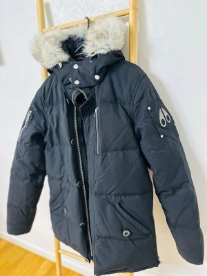 Svart pufferjacka Moose Knuckles S - Säljer en svart pufferjacka från Moose Knuckles med fet pälskant på huvan och coola metalldetaljer på ärmen. Jackan har dragkedjor vid ärmslut och flera fickor med dragkedja framtill. Perfekt för kalla dagar och riktigt snygg streetstil.