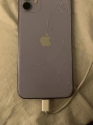 iPhone 11 - Lila iPhone 11 med synliga sprickor på baksidan. Telefonen är i fungerande skick och laddas via kabel. Perfekt för dig som vill ha en prisvärd smartphone trots kosmetiska defekter. Kameran och Apple-loggan är tydliga, men baksidan har flera sprickor.