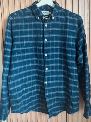 Blå rutig skjorta från East West - Säljer en blå rutig skjorta från East West med klassisk krage och vita knappar framtill. Skjortan har lång ärm och är tillverkad i mjuk bomull, perfekt för en avslappnad men snygg look.