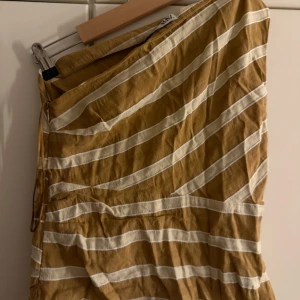 Randig oneshoulder topp i linne - Säljer en snygg oneshoulder topp från Zara i brunt och vitt med breda diagonala ränder. Toppen är ärmlös och har en cool asymmetrisk form. Materialet är linne vilket gör den lätt och skön att bära. Perfekt för varma dagar och sommarhäng.