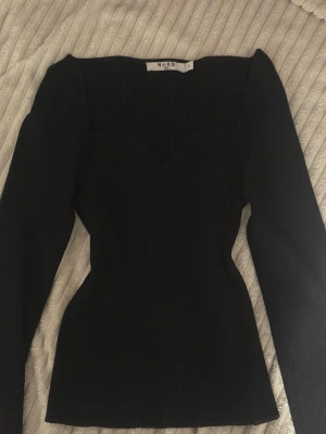 Svart långärmad topp från NA-KD - Säljer en svart långärmad topp från NA-KD med figurnära passform och snygg sweetheart-ringning. Toppen har en ribbad struktur och är tillverkad i ett mjukt syntetmaterial. Perfekt att styla med jeans eller kjol för en trendig look.