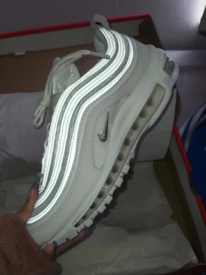 Nike Air Max 97 i vitt - Säljer ett par Nike Air Max 97 sneakers i vitt med reflekterande detaljer. Skorna har den ikoniska vågiga designen, synlig Air-sula och meshpartier för extra andningsförmåga. Swoosh-logga på sidan och snörning framtill. Perfekta för dig som vill sticka ut. Helt nya och oanvända, kommer med boxen.