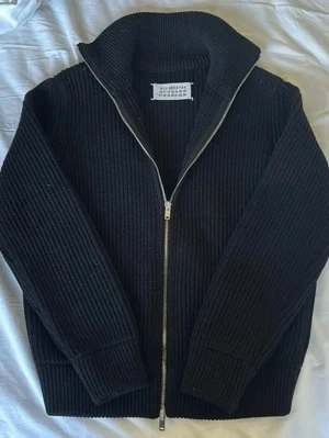 Svart stickad kofta från Maison Margiela - FAST PRIS! Säljer en svart, grovstickad kofta från Maison Margiela med hel dragkedja framtill och hög krage. Tröjan har långärmad passform och är tillverkad i ull, vilket gör den perfekt för kyliga dagar. Diskret märkning i nacken med Margielas signaturstygn.