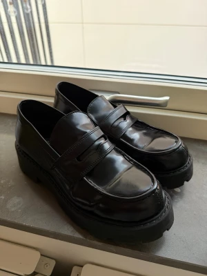 Svarta chunky loafers från Vagabond - Säljer ett par svarta loafers från Vagabond med chunky platt sula och rund tå. Skorna är tillverkade i blankt skinn och har klassisk pennyloafers-design. Perfekta för dig som gillar stilrena och trendiga skor med lite extra höjd.