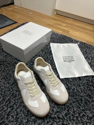 Maison Margiela vita sneakers med beige detaljer - Maison Margiela sneakers i vitt och beige med klassisk design. Skorna har en platt gummisula, snörning och paneler i mocka och skinn. Kommer med originalkartong och dustbag. Perfekt för dig som gillar stilrena och lyxiga sneakers.