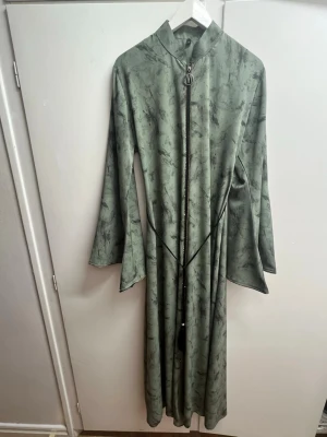 Grön långklänning med dragkedja - Snygg långklänning i grön nyans med svart marmorliknande mönster. Klänningen har lång ärm, hög krage och en dekorativ dragkedja framtill. Svart knytband i midjan med tofsar ger extra stil. Materialet känns mjukt och har ett fint fall. Väldigt modest och passar perfekt till vardagen men också till event, perfekt för dig som bär slöja, den har en lite vid passform och kan enkelt justeras med hjälp av bandet, pris går alltid att diskuteras💕