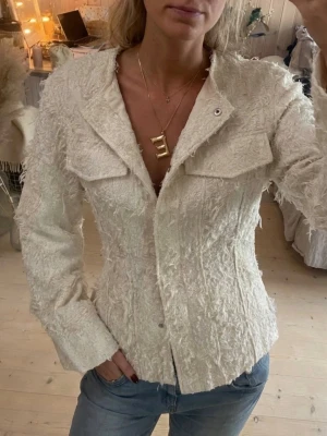 Offwhite Bree jacka från STOCKH LM Studio - Säljer en offwhite Bree jacka från STOCKH LM Studio i storlek XS. Jackan har en unik, ruffad textur och raka, långa ärmar. Den är figursydd med två bröstfickor och knappar framtill. Perfekt för dig som gillar trendiga detaljer och stilren look.
