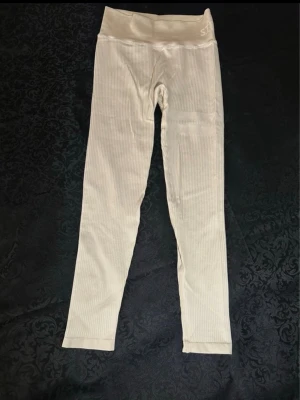 Vita ribbade leggings från Stronger S - Snygga vita leggings från Stronger med ribbad struktur och hög midja. De är stretchiga och har en diskret logga på linningen. Perfekta för träning eller chill, med en tight passform som sitter skönt. Materialet är mjukt och följsamt.