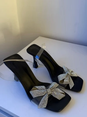 Svarta sandaler med glittrig strassrosett - Säljer ett par svarta sandaler med transparenta remmar och stora glittriga rosetter i strass framtill. Storlek 37, kan passa 36. Skorna har fyrkantig tå och lagom hög klack, perfekta för dig som vill sticka ut. Materialet är syntet och detaljerna gör dem riktigt trendiga. Riktigt passande classy stil, älskar dem men tyvärr för små men kommer köpa i rätt storlek. 