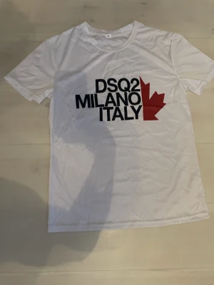 Vit DSQ2 Milano Italy t-shirt - Snygg vit t-shirt från DSQ2 med svart text 'MILANO ITALY' och en röd lönnlövsgrafik på bröstet. Klassisk passform och rund halsringning, perfekt till jeans eller shorts. Materialet känns mjukt och skönt mot huden.