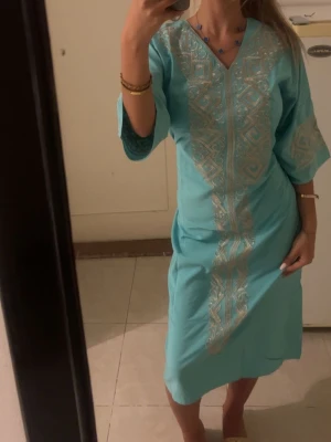 Turkos klänning med guldbroderi - Säljer en turkos klänning med v-ringning och vackra guldfärgade broderier framtill och på ärmarna. Klänningen har trekvartsärm och är knälång, perfekt för dig som gillar färg och detaljer. Materialet känns lätt och bekvämt. Den köptes utomlands och är 1 exemplar super bekväm och i storlek S